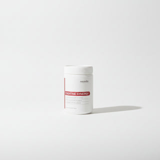 A white tub of 'monvita' Creatine Synergy powder sits next to a glass of vibrant pinkish-red creatine solution and a small metal spoon on a terracotta-colored surface, with a modern silver table lamp in the background.