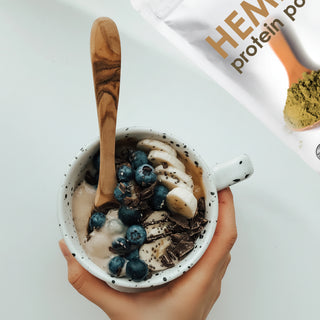 A hand holds a white speckled mug filled with protein powder, topped with blueberries, banana slices, and chocolate shavings, with a wooden spoon inside. A white pouch of hemp protein powder is visible in the top right corner on a clean white background.