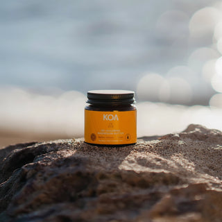 A small amber jar with a black lid and a bright orange label reading KOA, placed on a rough rock with a blurred beach or water background.