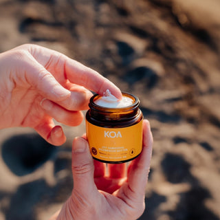 A person's hands holding an amber glass jar of KOA Joy Hormonal Magnesium Butter, with a dollop of white cream on the lid, set against a blurred sandy beach background.