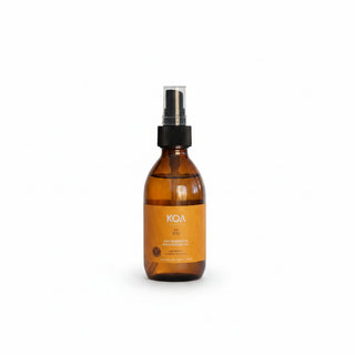 An amber glass spray bottle with a black pump and clear cap stands on a rock outdoors, with a blurred beach or ocean background. The label shows the brand KOA.