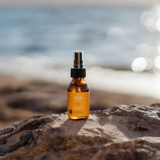 An amber glass spray bottle with a black pump and silver nozzle, labeled KOA, standing on a rocky beach with a blurred ocean background.
