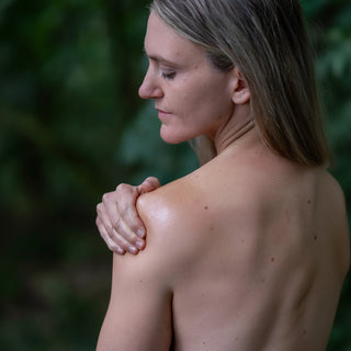 A close-up side profile of a woman with her back turned, holding her shoulder, set against a soft, blurred dark green background.