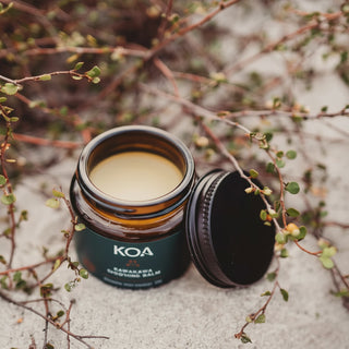 An open amber glass jar of KOA Kawawaka Healing Balm with its black lid resting beside it, styled on a sandy surface surrounded by small green leaves and twigs.